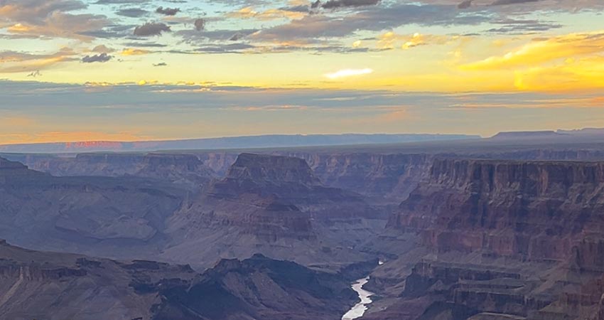 The golden hour casts a yellow and purple hue over the Grand Canyon and Colorado River at sunset, seen from the Desert View Point along the South Rim.