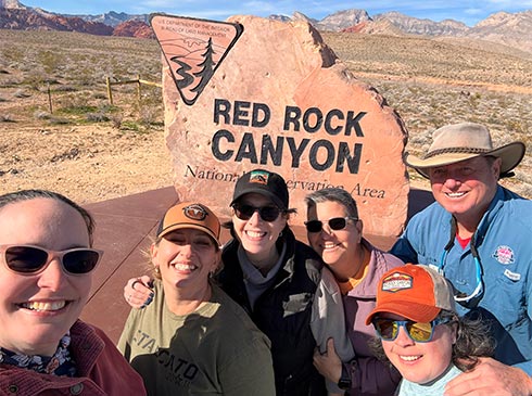 Pink Jeep Tours adventure guide and five female guests posing by the Red Rock Canyon National Conservation Area stone-cut sign.
