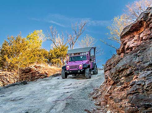 Pink Jeep descending steep, rocky trail alongside Baird Mountain fall foliage and a deep blue sky, Branson, MO.