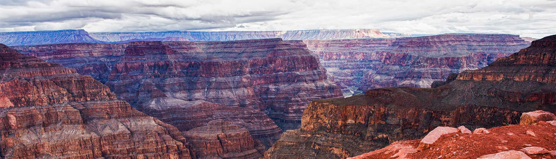 Panoramic landscape of the rust colored, multi-hued rock layers of the Grand Canyon's West Rim with the Colorado River below.
