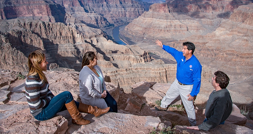 Pink Jeep Tours adventure guide talking to guests seated at the edge of the Grand Canyon West Rim and gesturing to the Colorado River below.
