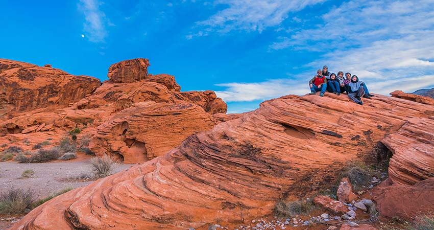 Family seated at the top of cow pie rock formation surrounded by Valley of Fire's red rock landscape, with a tiny moon peeking through the blue sky.