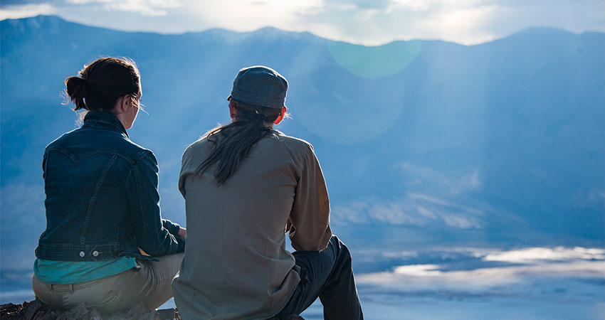 Sunbeams stream down on a couple sitting atop the Hell's Gate viewpoint overlooking the salt flats of Death Valley National Park.