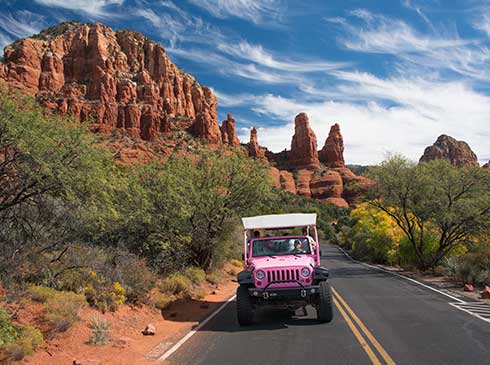 Pink Jeep Wangler driving towards the camera along a scenic road near Chapel of Holy Cross, with Sedona's red rock in the background.
