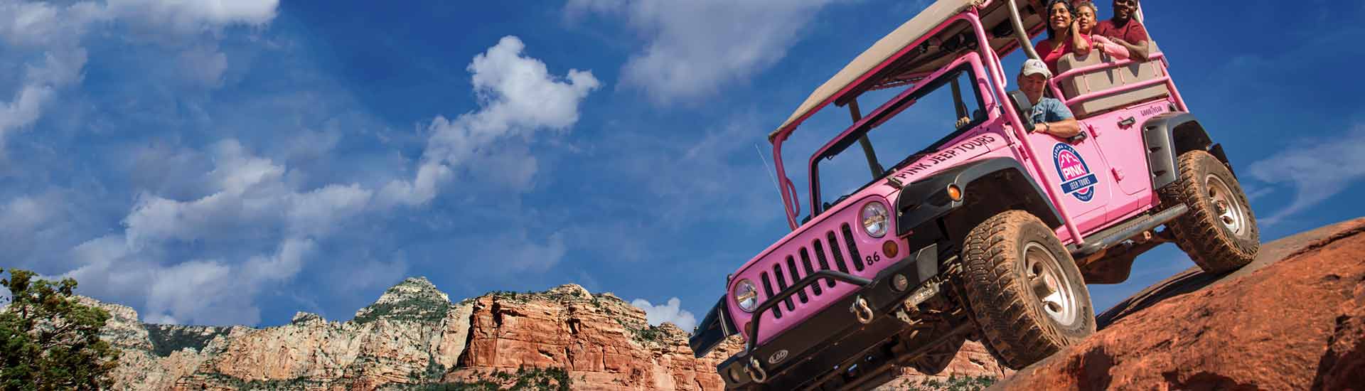 Pink Jeep Wrangler with passengers is perched atop a red rock cliff against a bright blue sky, Pink Jeep Tours Sedona.