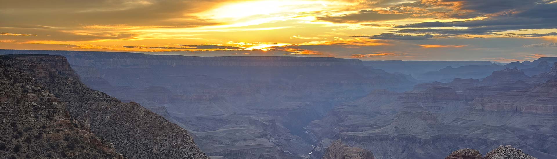 The rugged Grand Canyon landscape painted in hues of purple light under a vibrant yellow sunset, seen from Desert View Point along the South Rim.