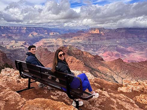 Man and woman sitting on a park bench perched on a rocky ledge overlooking the Grand Canyon, looking back at the camera with puffy clouds above.