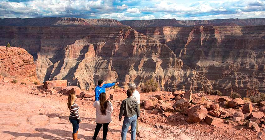 Four tour guests standing with a Pink Adventure Tours' guide at the edge of the Grand Canyon West Rim as he points towards the canyon's multicolored rock layers.